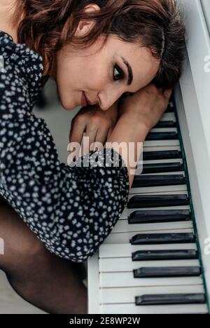 A beautiful brunette in a black dress put her head on the keyboard of a white piano Stock Photo