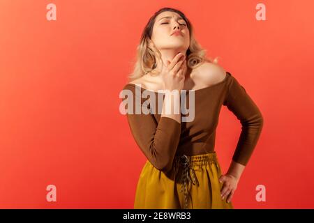 POrtrait of sad woman with blonde hair and closed eyes palping neck feeling throat ache and pain during swallow, tonsillitis inflammation. Indoor stud Stock Photo