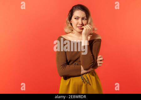 Oh no! Portrait of young nervous woman with blonde hair in brown blouse, thinking and worried about work, deadlines and biting her nails. Indoor studi Stock Photo