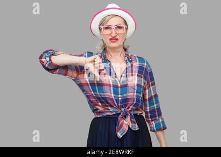 Dislike. Portrait of sad dissatisfied modern stylish mature woman in casual style with hat and eyeglasses standing looking at camera with thumb down. Stock Photo