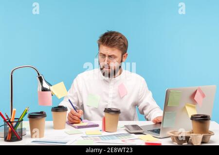 Busy concentrated man office worker sitting at workplace all covered with sticky notes and writing down thoughts in notepad, drinking coffee. Indoor s Stock Photo