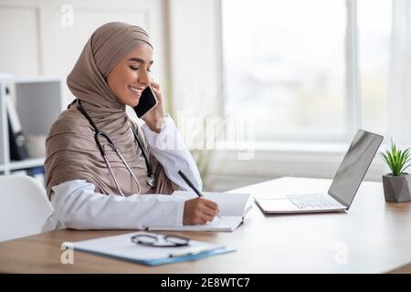 Lady in hijab doctor talking on phone and taking notes Stock Photo