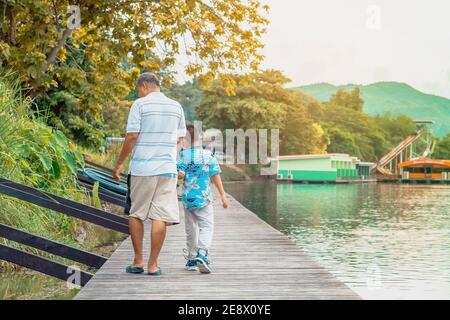 Back view of Asian grandfather and grandchild wear protective face mask to prevent Coronavirus (COVID-19) walking in a nature path on wooden bridge al Stock Photo