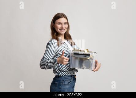 Smiling Nature-Friendly Woman Holding Container With Metal Waste And Showing Thumb Up Stock Photo