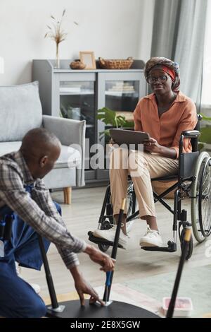 Vertical portrait of young African-American woman in wheelchair watching handyman assembling furniture, online service and assistance concept Stock Photo