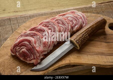 Several slices of raw, rolled British lamb displayed on an olive wood chopping board with a homemade knife. England UK GB Stock Photo