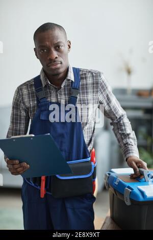 Vertical portrait of African-American handyman looking at camera while standing with toolbox in home interior Stock Photo