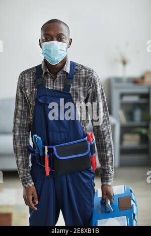 Vertical portrait of African-American handyman wearing mask and holding toolbox while standing in home interior looking at camera Stock Photo