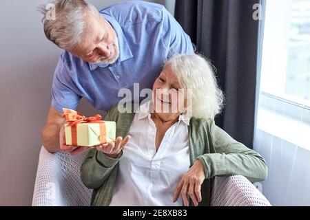 senior female person with satisfied face getting present box by her nic handsome husband, gray-haired elderly couple celebrating woman's birthday, man Stock Photo