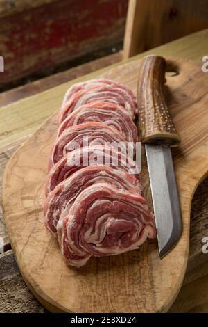 Several slices of raw, rolled  British lamb displayed on an olive wood chopping board with a homemade knife. England UK GB Stock Photo