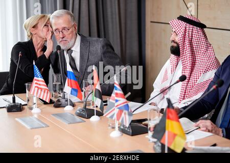 caucasian delegates sharing opinions about colleague speech, adult female talk something in the ear of senior gray bearded man in suit, discussing whi Stock Photo