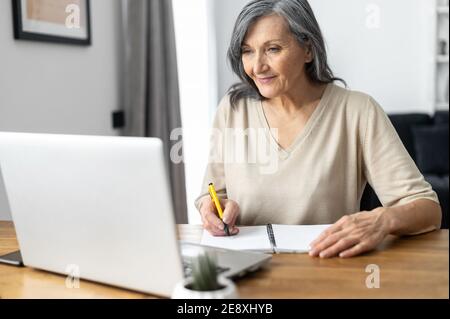 Charming mature mid-aged woman watching online webinars on the laptop sitting at the table at home, taking notes, e-learning. Senior business lady Stock Photo
