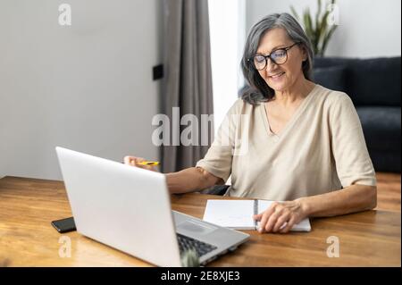 Mature focused woman watching online webinars on the laptop sitting at the table at home, taking notes, e-learning. Concentrated senior business woman Stock Photo