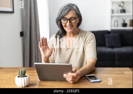 Cheerful confident senior woman using a digital tablet for video connection, remote meeting, smiling mature lady waving hello into webcam, greeting Stock Photo