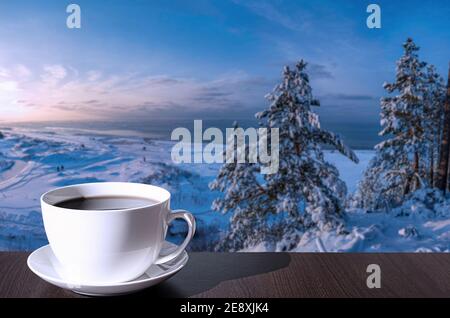 Cup of coffee on the table with view of snowy beach with dunes, Baltic sea and covered in snow fir trees on the hill. Stock Photo