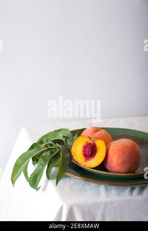 Peach in halves with bone in minimalistic style. Peaches with leaves on color green plate on table with white tablecloth. Ripe juicy peaches Still Stock Photo
