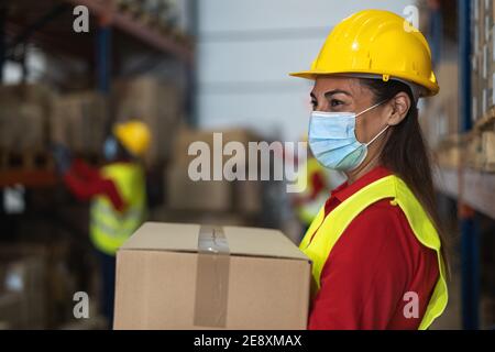 Latin woman working in warehouse loading delivery boxes while wearing face mask during corona virus pandemic - Logistic and industry concept Stock Photo