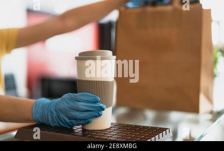 Bar owner working only with take away orders during corona virus outbreak - Young woman worker wearing surgical gloves giving takeout meal and coffee Stock Photo