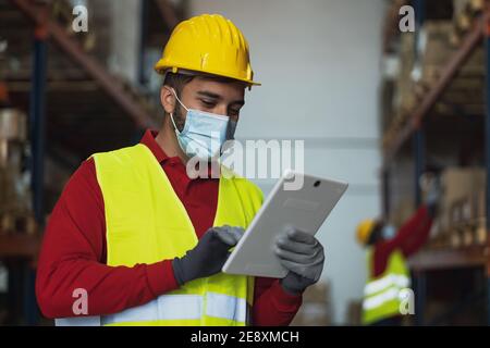 Young man working in warehouse doing inventory using digital tablet and loading delivery boxes plan while wearing face mask during corona virus Stock Photo