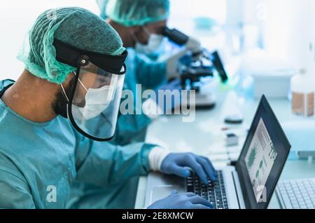 Doctors wearing personal protective equipment working in laboratory using computer and examining coronavirus through microscope Stock Photo