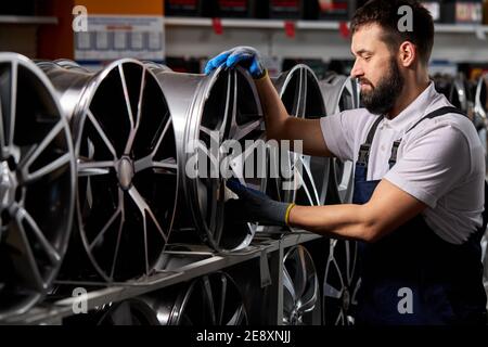 male auto mechanic checking new tires and discs in auto shop, preparing for sale, automobiles industry, bearded male in uniform at work place Stock Photo