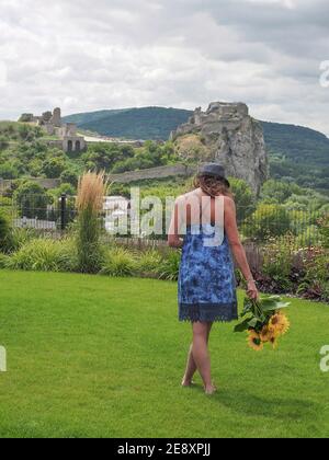 Woman in summer dress and hat with sunflowers in hand walking in garden. Devin Castle on background. Stock Photo