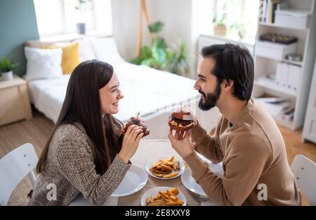 Young couple in love eating hamburgers indoors at home.