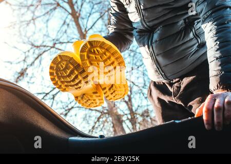 A man takes a pair of yellow rubber boots out of the trunk for a walk through the mud and snow - a country trip by car to pick mushrooms in the forest Stock Photo