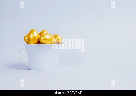 Decorative Easter eggs are painted gold and stacked in a blue porcelain cup on a light blue background. Easter, spring concept. Selective focus. Stock Photo