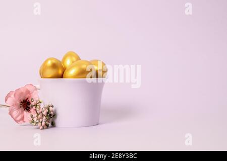 Easter composition of golden decorative eggs in a porcelain cup and a flower on a pink background. Selective focus, copy space. Stock Photo