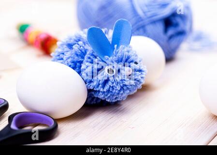 This blue rabbit is made of pompons for Easter decor by hand. Easter bunny and white eggs on a stand Children's art project. DIY concept. Stock Photo