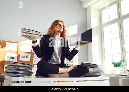 Calm worker holding papers and laptop and meditating in yoga pose on office desk Stock Photo