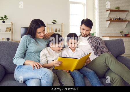 Parents with little daughter and son read fairy tales in a book sitting on the couch at home. Stock Photo