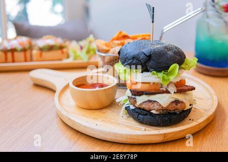 Black burger with meat patty, cheese, tomatoes, mayonnaise, french fries in a paper cup and glass of cold cola soda with ice. Dark wooden rustic table Stock Photo