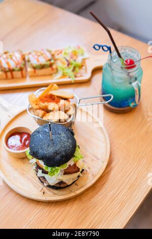 Black burger with meat patty, cheese, tomatoes, mayonnaise, french fries in a paper cup and glass of cold cola soda with ice. Dark wooden rustic table Stock Photo