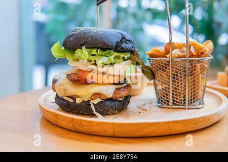 Black burger with meat patty, cheese, tomatoes, mayonnaise, french fries in a paper cup and glass of cold cola soda with ice. Dark wooden rustic table Stock Photo