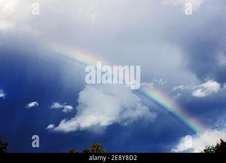 Real pretty rainbow in dark sky through white clouds summer day Stock Photo