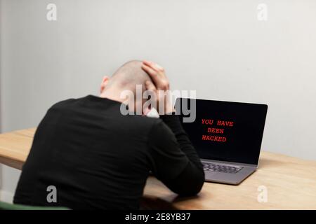 Red text on a laptop saying ‘You have been hacked’. A desperate man sitting in front of his infected computer. Cybercrime, infected unsafe device Stock Photo