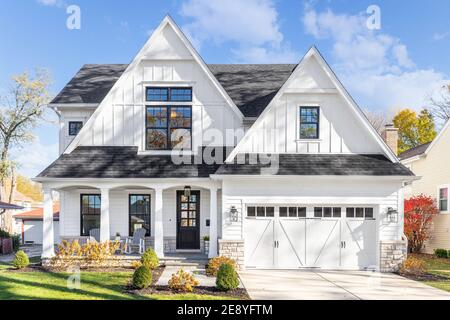 A new, white modern farmhouse with a dark shingled roof and black window frames. The bottom of the house has a light rock siding and front porch. Stock Photo