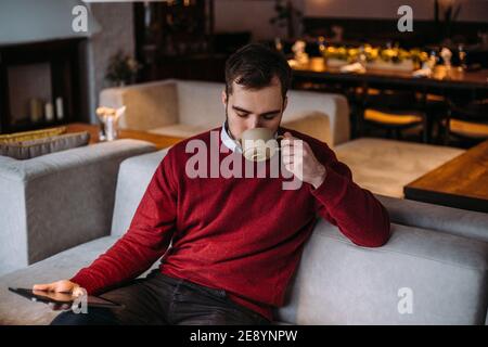 A young handsome man sits in a cafe with a cup of coffee and looks into a gadget. The concept of working in a cafe Stock Photo