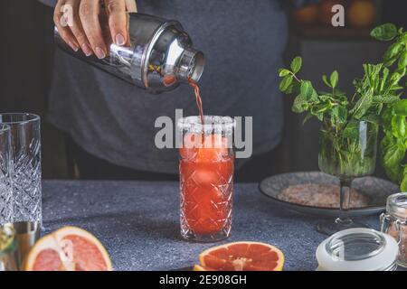 Real woman preparing pink Grapefruit Mezcal Paloma Cocktail in highball glass with water drops on table in real home. New life concept of quarantine. Stock Photo