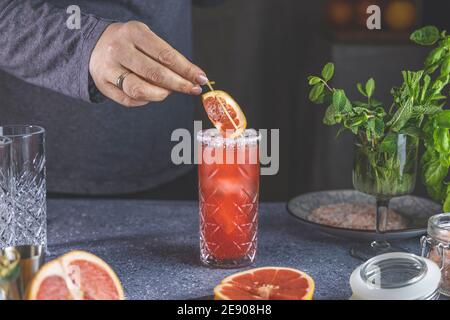 Real woman preparing pink Grapefruit Mezcal Paloma Cocktail in highball glass with water drops on table in real home. New life concept of quarantine. Stock Photo