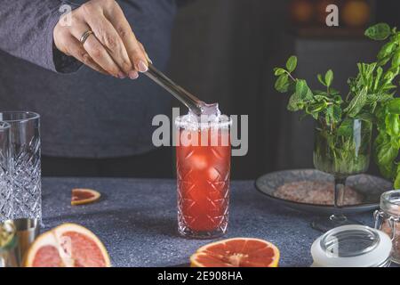 Real woman preparing pink Grapefruit Mezcal Paloma Cocktail in highball glass with water drops on table in real home. New life concept of quarantine. Stock Photo