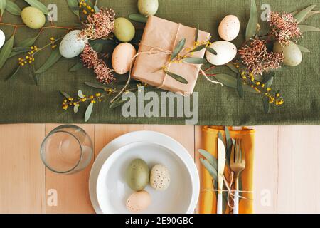 White wooden table with white plate and bowl, pastel colored Easter eggs, fresh garden flowers and brown paper wrapped gift, green table runner Stock Photo