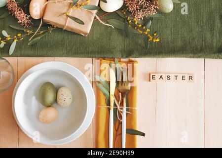 White wooden table with white plate and bowl, pastel colored Easter eggs, fresh garden flowers and brown paper wrapped gift, green table runner Stock Photo