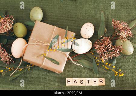 White wooden table with white plate and bowl, pastel colored Easter eggs, fresh garden flowers and brown paper wrapped gift, green table runner Stock Photo
