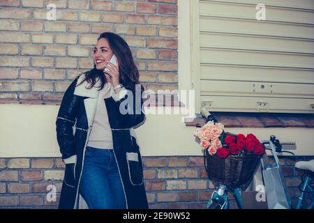 Beautiful young woman chatting with her friend on mobile phone, leaning against a brick wall in the evening while waiting someone. Stock Photo