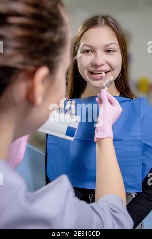 Close up portrait of beautiful young lady sitting in dental chair while stomatologist hands in sterile gloves holding tooth samples. Stock Photo