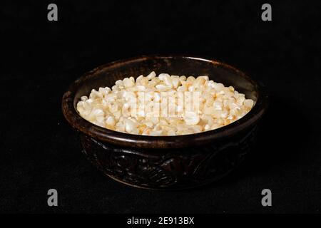 Broken white corn seeds in a wooden jar on a black surface. Stock Photo