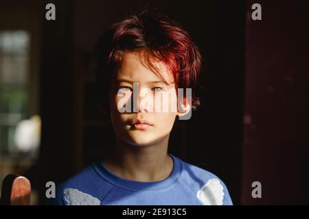 A boy with dyed red hair stands in dark room in bright patch of light Stock Photo